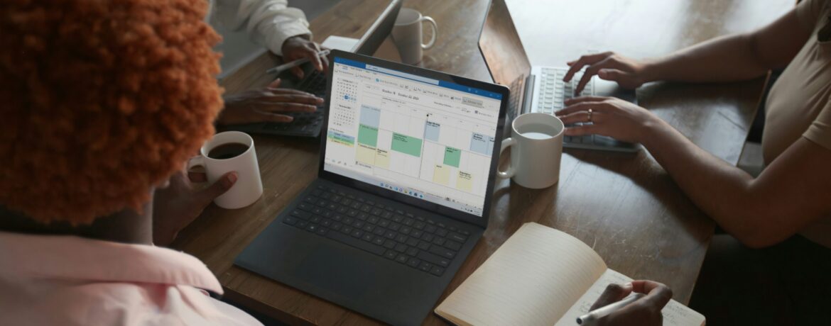 Three people in a meeting at a table discussing schedule on their Microsoft laptop